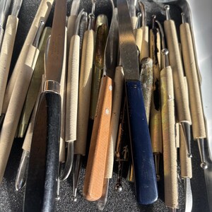 May include: A collection of various sculpting tools, including knives and rounded tip tools, with wooden and dark blue handles. The tools are arranged in a metal tray with a dark gray foam insert, showcasing a variety of shapes and sizes.