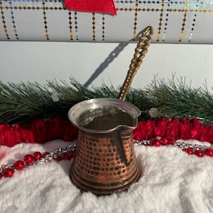 May include: A copper-colored Turkish coffee pot with a hammered texture and a decorative handle. The pot sits on a white, fluffy surface, with red and silver decorative elements in the background. The pot is approximately 4 inches tall.