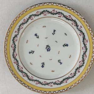 May include: A white porcelain plate with a yellow border and gold trim. The plate features a floral design with blue flowers and green leaves. The rim has a decorative pattern of red and green foliage.