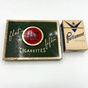 May include: Vintage Lucky Strike and Parliament cigarette packs. The Lucky Strike tin is green with a red and white circular logo and the words "flat fifties cigarettes". The Parliament pack is tan with blue accents and the brand name in elegant script.