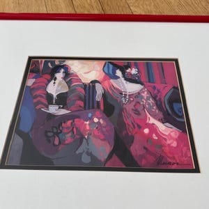 May include: A framed print featuring a stylized illustration of two figures in elegant attire. The artwork uses a palette of pinks, reds, and blues, with one figure holding a teacup. The print is set against a white mat and framed in red.