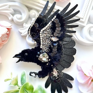 May include: A black bird brooch featuring intricate beadwork, sequins, and rhinestones. The bird is depicted in flight with wings spread, crafted from textured black material, dark feathers, and sparkling crystal accents on the body and wings.