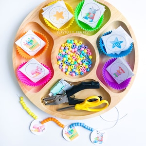 May include: A wooden tray with colorful cupcake liners filled with paper circles featuring animal illustrations. The tray also includes a pile of colorful beads, a hole punch, scissors, and a string of beads with paper circles attached.