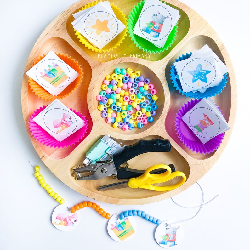 May include: A wooden tray with colorful cupcake liners filled with paper circles featuring animal illustrations. The tray also includes a pile of colorful beads, a hole punch, scissors, and a string of beads with paper circles attached.