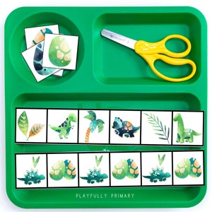 Puede incluir: Una bandeja de pl&aacute;stico verde con dos filas de tarjetas de im&aacute;genes con tem&aacute;tica de dinosaurios. La fila superior muestra una palmera, un dinosaurio, una hoja y un huevo de dinosaurio. La fila inferior muestra un huevo de dinosaurio, un dinosaurio, un huevo de dinosaurio y un dinosaurio. La bandeja tambi&eacute;n incluye un par de tijeras amarillas y un juego separado de tarjetas de huevos de dinosaurio.