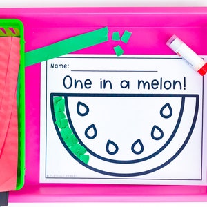 May include: A pink tray holds a green basket with red and green paper strips, a glue stick, and a worksheet. The worksheet features a watermelon outline with the text "One in a melon!" and space for a name.
