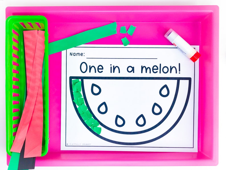 May include: A pink tray holds a green basket with red and green paper strips, a glue stick, and a worksheet. The worksheet features a watermelon outline with the text "One in a melon!" and space for a name.