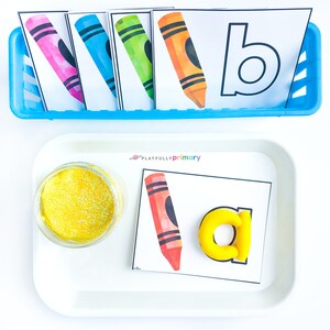 May include: A white tray with a small jar of yellow glitter, a red crayon, and a white card with the letter 'a' written on it. The letter 'a' is made of yellow playdough. There is a blue plastic bin with cards showing different colored crayons and the letter 'b' in the background.