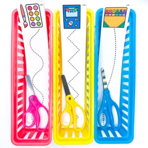 May include: Three colorful plastic storage bins with a wavy, zig-zag, and straight line pattern on white paper inside. Each bin has a pair of child-sized scissors and a picture at the top. The pictures are of a paint palette, a rocket ship, and crayons.