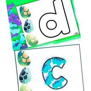 Puede incluir: Dos tarjetas blancas con contornos negros de las letras 'D' y 'C'. La letra 'D' est&aacute; en una tarjeta verde con un borde. La letra 'C' est&aacute; en una tarjeta blanca. Ambas tarjetas tienen huevos de dinosaurio coloridos y figuritas de dinosaurios azules.