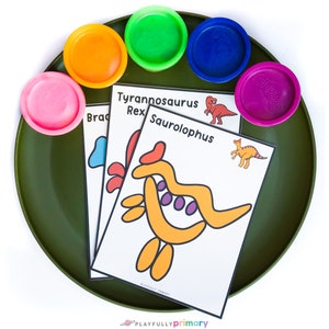 Puede incluir: Un plato verde con cuatro recipientes de plastilina de colores y tres tarjetas de dinosaurios. Las tarjetas tienen los nombres "Tyrannosaurus Rex", "Saurlophus" y "Brachiosaurus" en ellas. Las tarjetas tambi&eacute;n tienen una imagen de cada dinosaurio.