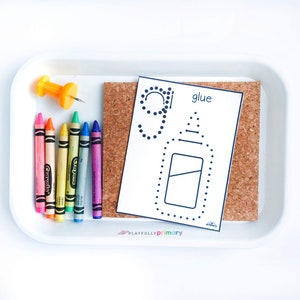May include: A white card with a dotted line drawing of a glue bottle and the word "glue" printed on it. The card is on a cork board. There are six crayons in a rainbow of colors on a white tray.