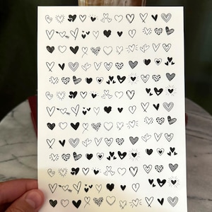 May include: A sheet of white paper with a variety of black heart-shaped stickers. The hearts are drawn in different styles, including simple outlines, filled-in shapes, and hearts with swirls and other details.