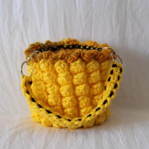 May include: A handmade yellow crochet bag with a textured, bubble-like pattern. The bag features a black and yellow strap and silver metal rings. The top edge has a darker, textured trim. The bag is displayed on a white background.