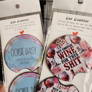 May include: Two car coasters in plastic packaging. One coaster has a blue floral design with the text "OOPSIE DAISY LOOKS LIKE YOU THOUGHT I GAVE A FUCK". The other has a wine glass pattern with the text "NOT ENOUGH WINE FOR THIS SHIT".