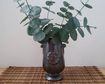 Può includere: Un vaso in ceramica marrone scuro con un motivo floreale e due manici, che contiene un mazzo di foglie di eucalipto verdi.
