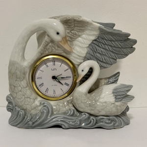 May include: A white porcelain clock shaped like two swans. The clock face is gold with black Roman numerals and black hands. The clock says "Alarm" below the clock face.