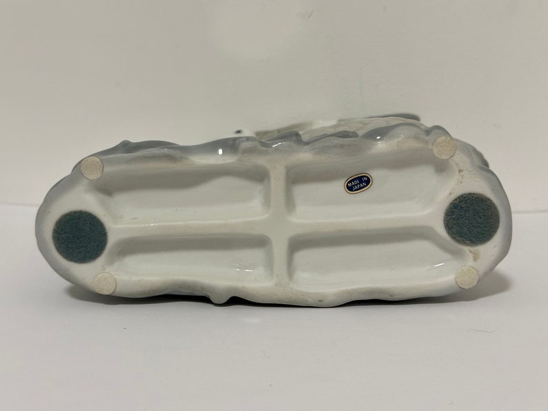 May include: White ceramic figurine with a gray glaze. The figurine has a detailed design and is marked "Made in Japan" on the bottom.