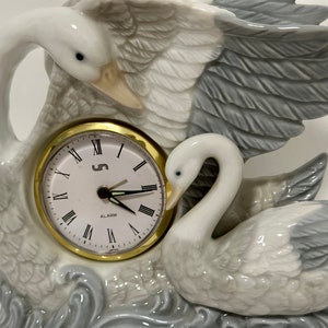 May include: A white porcelain swan clock with an alarm function. The clock face is white with black Roman numerals and black hands. The clock is set in a base with a gray and white swan design.