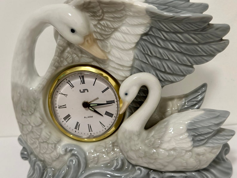 May include: A white porcelain swan clock with an alarm function. The clock face is white with black Roman numerals and black hands. The clock is set in a base with a gray and white swan design.