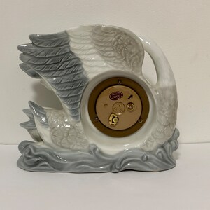 May include: A white porcelain swan clock with a gray and white feathered design. The clock face is visible and features a gold-colored rim and a small red label that reads "Swiss Made".