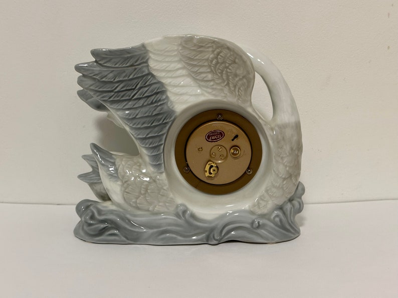 May include: A white porcelain swan clock with a gray and white feathered design. The clock face is visible and features a gold-colored rim and a small red label that reads "Swiss Made".