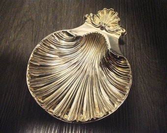 Könnte beinhalten: Eine silberfarbene, muschelförmige Schale mit detailliertem, geriffeltem Design. Die Schale hat einen dekorativen, verzierten Griff. Die Muschelschale wird wahrscheinlich zum Servieren oder Ausstellen verwendet.
