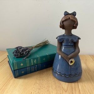 May include: A ceramic figurine of a girl wearing a blue dress with a white flower in her hand. The figurine is standing on a wooden surface with two books and a bunch of lavender.