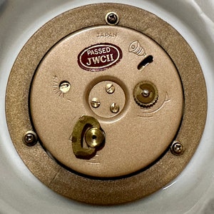 May include: Close-up of a gold-colored clock mechanism with a brown label that reads "PASSED JWCI" and "JAPAN".