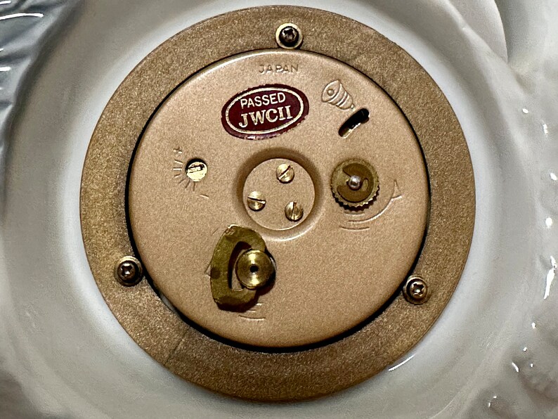 May include: Close-up of a gold-colored clock mechanism with a brown label that reads "PASSED JWCI" and "JAPAN".
