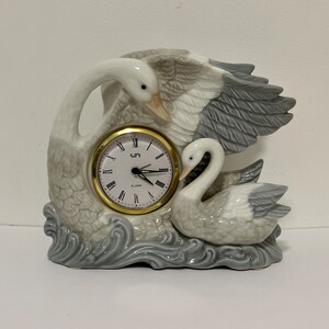 May include: A white and gray porcelain clock shaped like two swans. The clock has a gold-colored face with black Roman numeral markings and a black second hand. The clock is set on a base that looks like water.