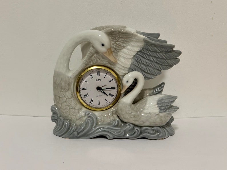 May include: A white and gray porcelain clock shaped like two swans. The clock has a gold-colored face with black Roman numeral markings and a black second hand. The clock is set on a base that looks like water.