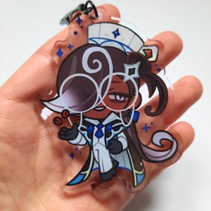Puede incluir: Un charm de acr&iacute;lico transparente con un personaje de dibujos animados con cabello casta&ntilde;o, que lleva un atuendo blanco y azul y gafas. El personaje sostiene una peque&ntilde;a taza de caf&eacute;.