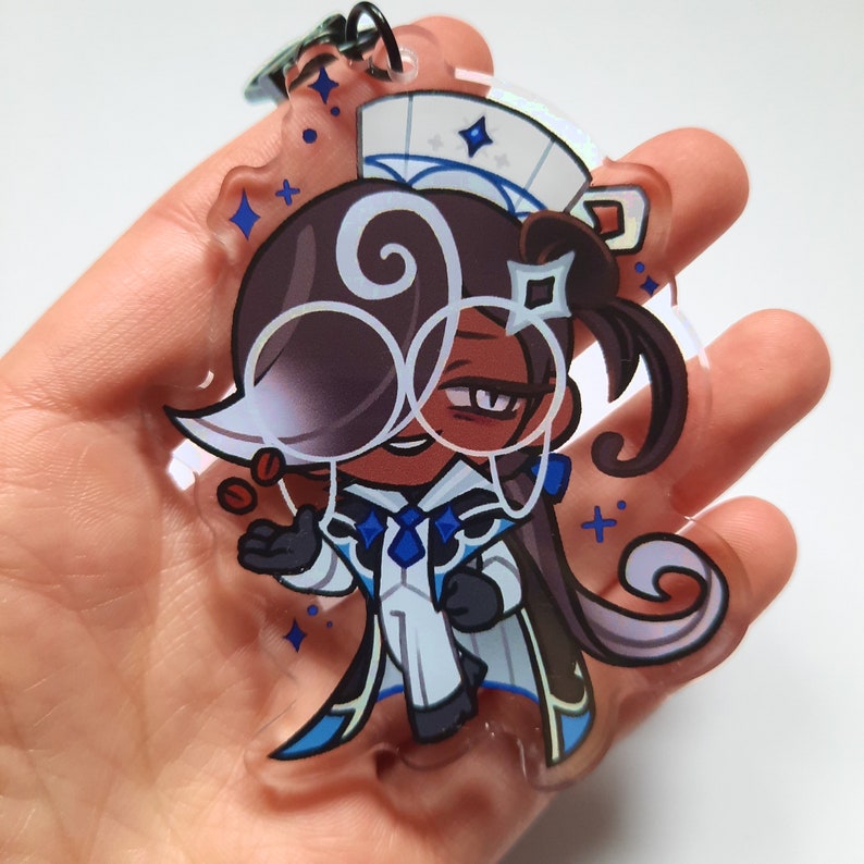 Puede incluir: Un charm de acr&iacute;lico transparente con un personaje de dibujos animados con cabello casta&ntilde;o, que lleva un atuendo blanco y azul y gafas. El personaje sostiene una peque&ntilde;a taza de caf&eacute;.