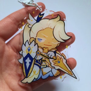 Puede incluir: Un llavero de acr&iacute;lico transparente con un personaje de dibujos animados de pelo rubio, vestido con un atuendo azul y dorado y sosteniendo una espada dorada. El personaje tiene una expresi&oacute;n seria y est&aacute; rodeado de estrellas doradas brillantes.