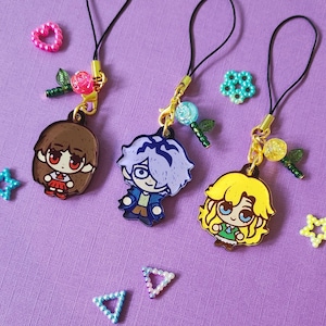 May include: Three anime-style character charms with black lanyards, gold-coloured clasps, and decorative beads. The charms feature a girl with brown hair, a boy with blue hair, and a girl with yellow hair. Additional decorative beads and charms are scattered around.