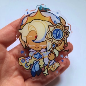 Puede incluir: Un amuleto de acr&iacute;lico transparente con un personaje de dibujos animados con cabello rubio, un atuendo estampado en azul y blanco y una corona dorada. El personaje sostiene un bast&oacute;n dorado con un ojo azul en la parte superior.