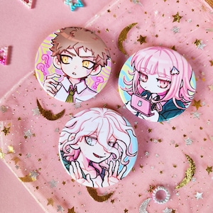 Può includere: Tre bottoni rotondi con spilla che raffigurano personaggi anime con capelli rosa, marroni e bianchi. I personaggi indossano abiti colorati e hanno espressioni diverse. I bottoni sono su uno sfondo rosa con stelle dorate e mezze lune.