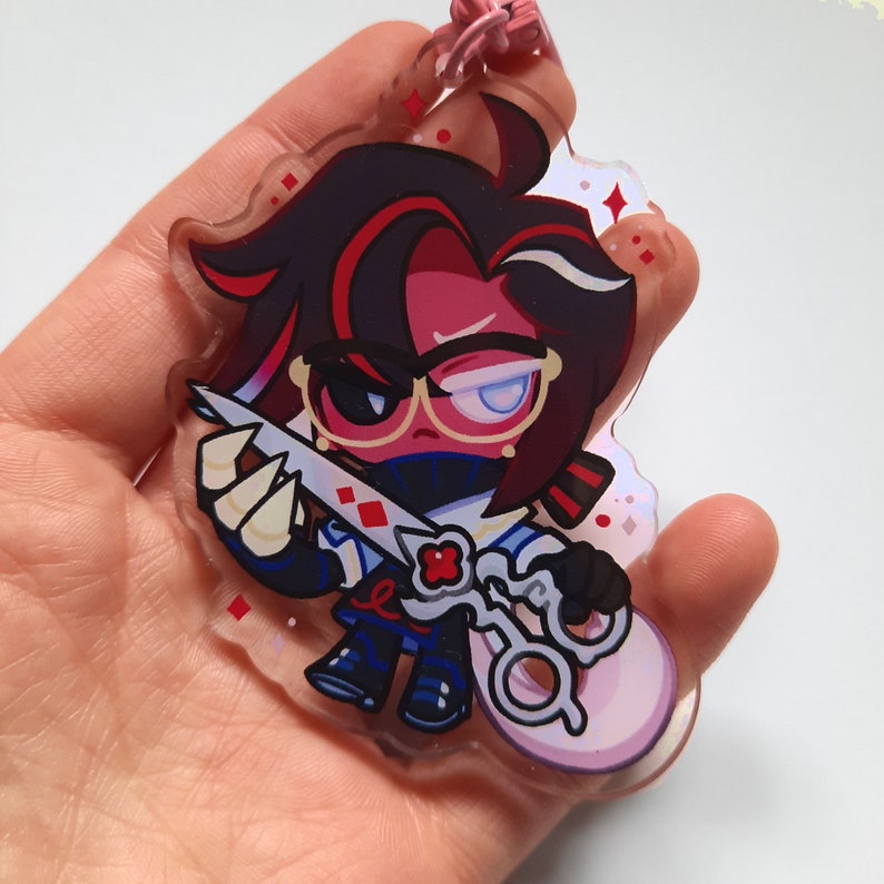 Puede incluir: Un llavero de acr&iacute;lico transparente con un personaje de dibujos animados con cabello rojo y negro, que lleva gafas y un atuendo blanco y azul. El personaje sostiene un par de tijeras y una flor roja.
