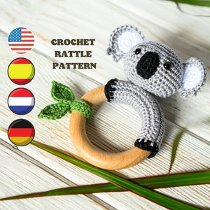 May include: Crochet koala rattle pattern with flags of the United States, Spain, Netherlands, and Germany. The rattle is made of a wooden ring with a crocheted koala attached. The koala is gray with black eyes and a black nose. There are two green crocheted leaves attached to the ring.