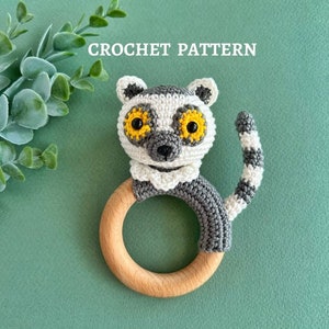 May include: Crochet pattern for a lemur rattle toy. The lemur is grey and white with yellow eyes and is attached to a wooden ring.