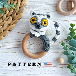 May include: A crocheted grey and white lemur toy with yellow eyes attached to a wooden teething ring. The toy is on a white wooden surface with a woven basket and green plants in the background. The text "PATTERN" is visible in the bottom left corner of the image.