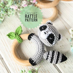 Peut inclure: Hochet en crochet en forme de raton laveur avec un anneau en bois et des feuilles vertes. Le hochet est gris, noir et blanc, avec le texte "CROCHET RATTLE PATTERN" sur un fond vert. Des fleurs ornent l'image.