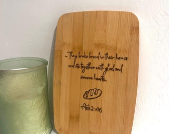 Puede incluir: Una tabla de cortar de madera con una cita sobre compartir el pan juntos con alegría y sinceridad. La cita es de Hechos 2:46.