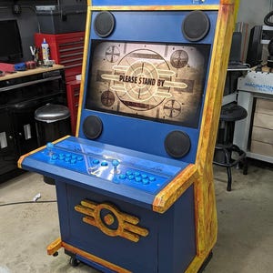 Puede incluir: Un gabinete de arcade azul y amarillo con el texto "VAULT-TEC" en la parte delantera. El gabinete tiene una pantalla que muestra el texto "PLEASE STAND BY".