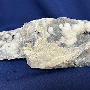 Könnte beinhalten: Ein rohes Mineralexemplar mit einer zerklüfteten, grauen und beigen Gesteinsmatrix, auf deren unebener Oberfläche kleine, weiße, kugelförmige Kristallformationen wachsen, was eine natürliche, unpolierte und strukturierte geologische Form zeigt.