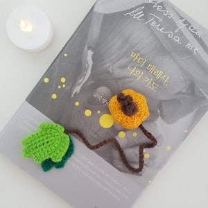 May include: A crocheted bookmark with a green leaf and a brown stem. The bookmark is attached to a crocheted orange pumpkin with a brown stem. The bookmark is on top of a book titled "Mother Teresa, My Prayer" with the text "Bless you less you Teresa me" on the cover.
