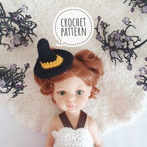 May include: A crocheted witch hat with a black brim and yellow crescent moon detail. The hat is worn by a doll with red hair.