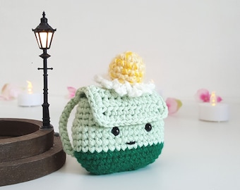 May include: A handmade crochet bag in shades of green, with a dark green base and a light green body. The bag features a small flap, black eyes, and a smiling mouth. A yellow and white crocheted flower sits on top. The bag has a handle.