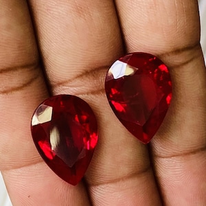 Op de afbeelding: Twee peervormige rode edelstenen, waarschijnlijk robijnen, worden op een hand getoond. De edelstenen zijn gefacetteerd en hebben een hoge glans.